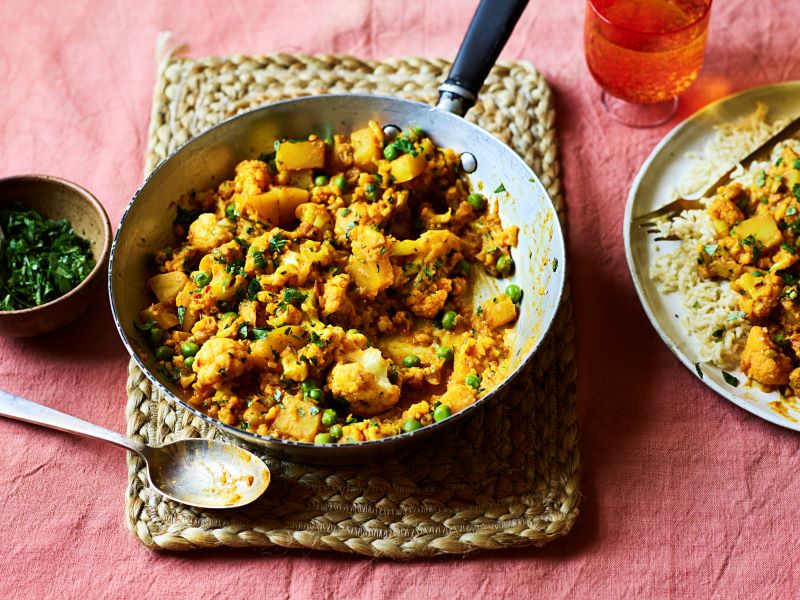 A frying pan with cauliflower, pea and potato curry, on a woven placemat on top of a pink table cloth and with a bowl of herbs.