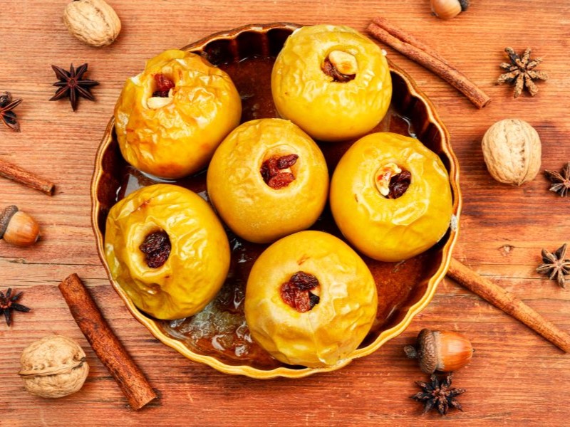 Top view of a tray of baked apples, stuffed with nuts and raisins on a wooden table that's decorated with cinnamon sticks, star anise, nutmeg and acorns.