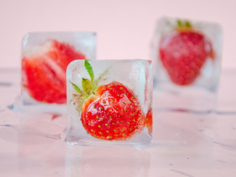 Close up of ice cubes with strawberries in, on a pale worktop and with a pink wall in the background.
