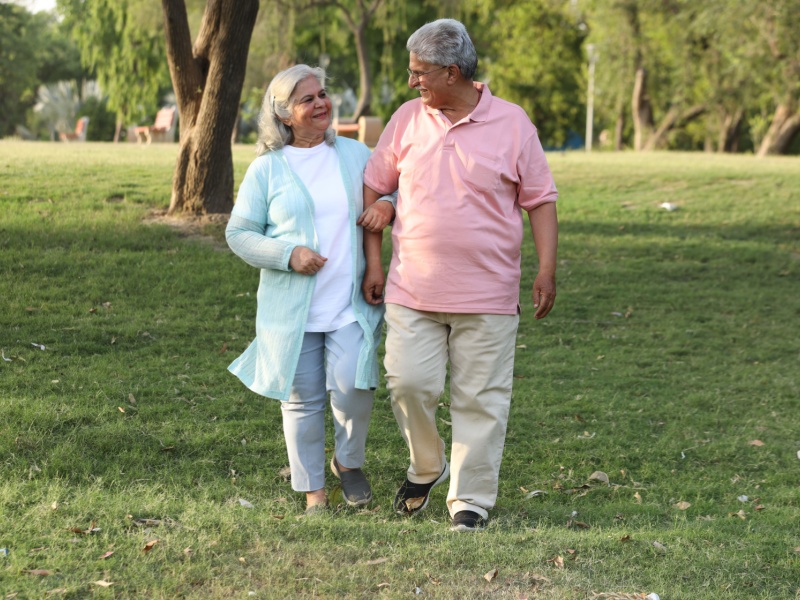 Senior South Asian couple walking through a park, with trees in the background, on a bright day.