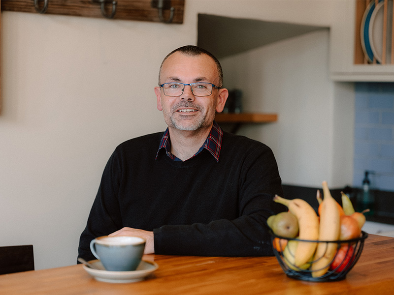 Ben Aitken sitting at a table with a hot drink and a fruit bowl.