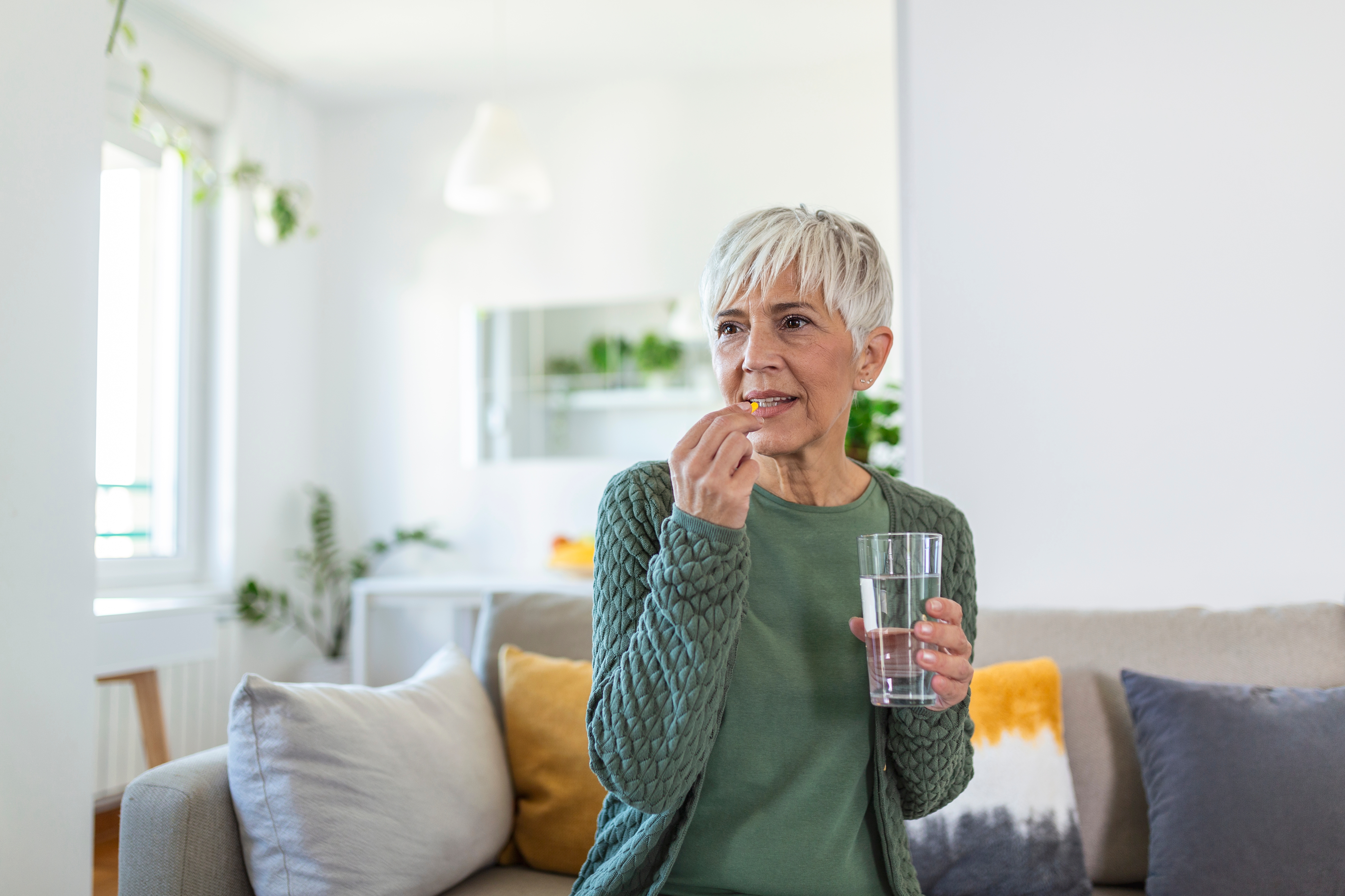 Older woman sitting on sofa taking pill with glass of water