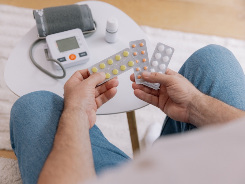 Close up, top view of a person sitting on the sofa holding packets of medicine and with a blood pressure monitor on a table in front of them.