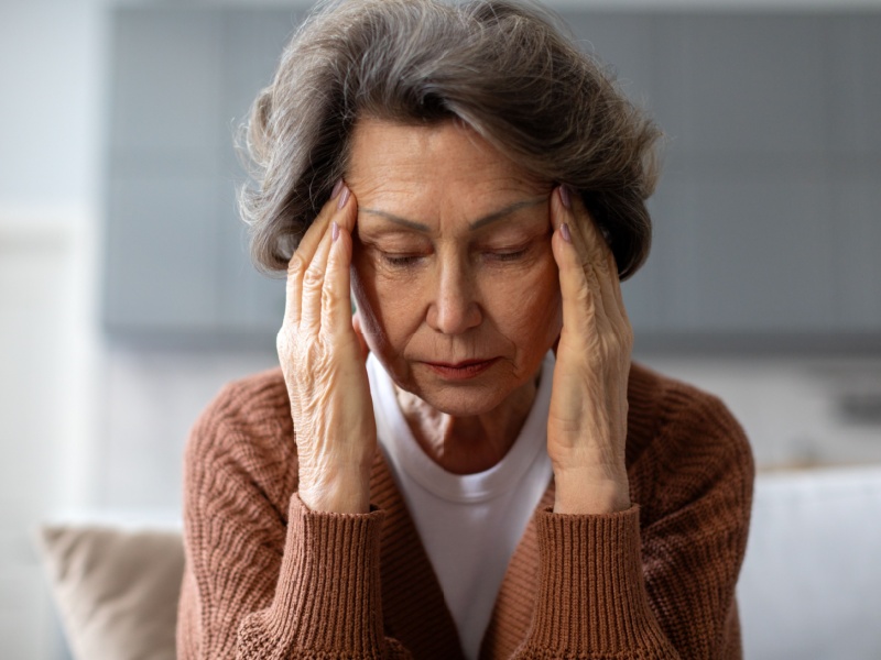 Close up of a senior woman with grey hair in a brown cardigan putting her hands to her temples and with her eyes closed