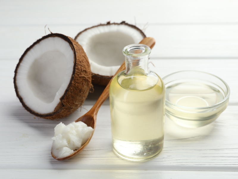 An opened coconut, a spoon with solid coconut oil on, and a clear glass bottle and small dish with liquid coconut oil. All on a white wooden background.