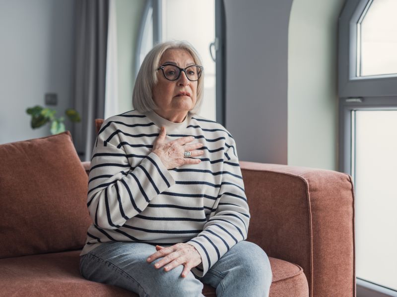 Older woman holding chest and looking worried while sitting on sofa