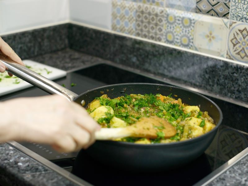 Close up of a person cooking chicken with herbs in a large rounded frying pan on an induction hob.