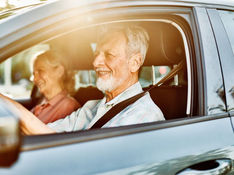 Close up of a senior man with grey hair and beard driving a car with the seat belt on and window open, a woman sits in the seat next to him.