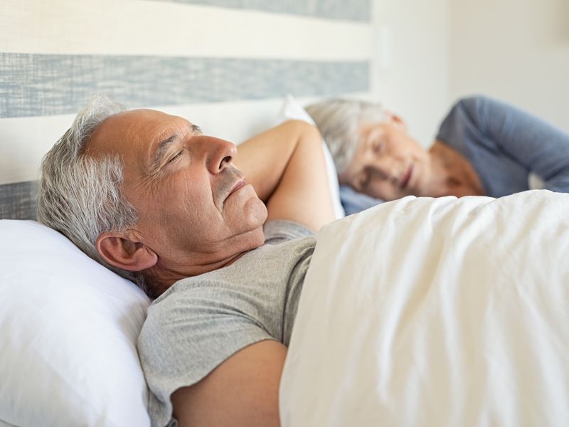 A senior couple lying in bed, the man in the foreground wearing a grey t-shirt and lying on his back.