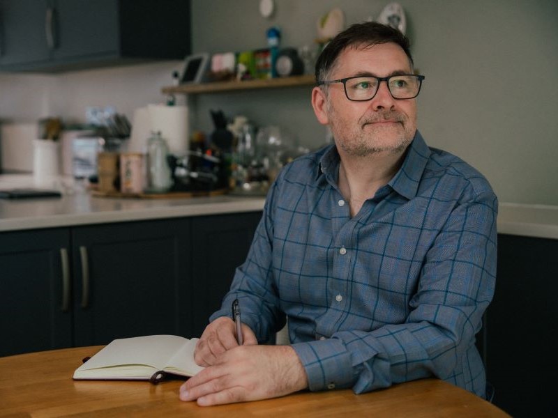 A middle-aged white man with dark hair and glasses is sitting at a kitchen table writing in a notebook. He is wearing a blue shirt and looking away from the camera.