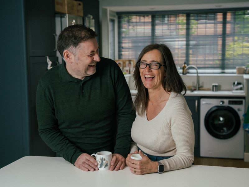 A middle-aged, dark-haired man in a dark blue jumper stands at a white kitchen worktop with a woman. She has long dark hair, glasses and a white v-neck top. They are laughing and holding cups of tea.