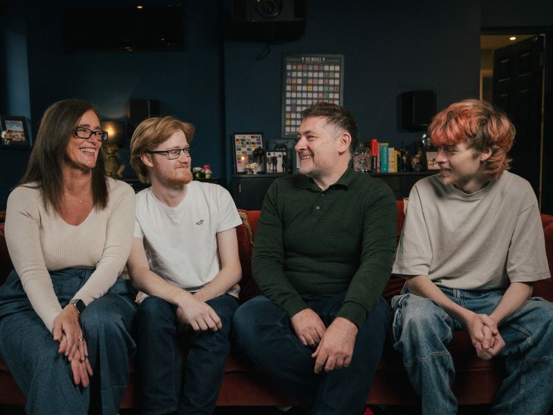A white middle-aged woman and man sit on a sofa in a row with their two young adult sons. The group are laughing together at a joke and all wearing jeans with plain tops.