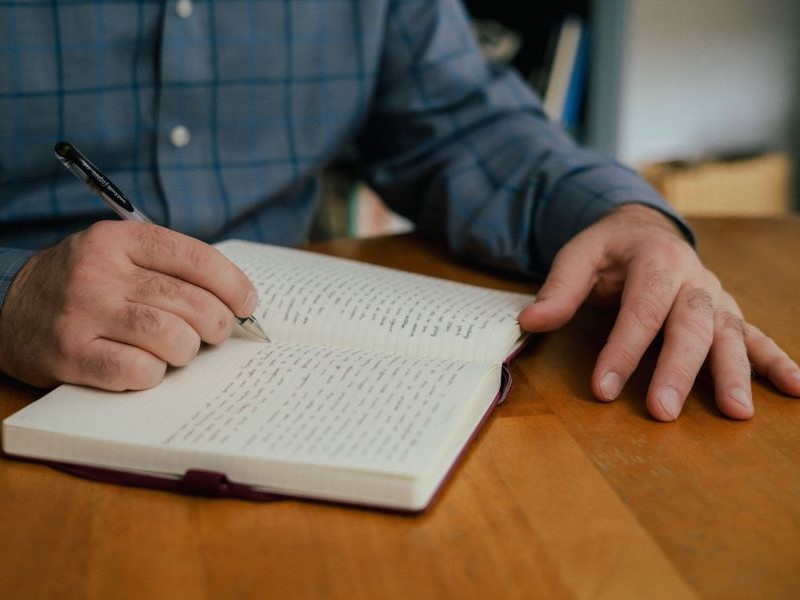 A close up of a white man's hands as he writes in a notebook. He is sat at a light wooden table and has a long-sleeved blue shirt on.