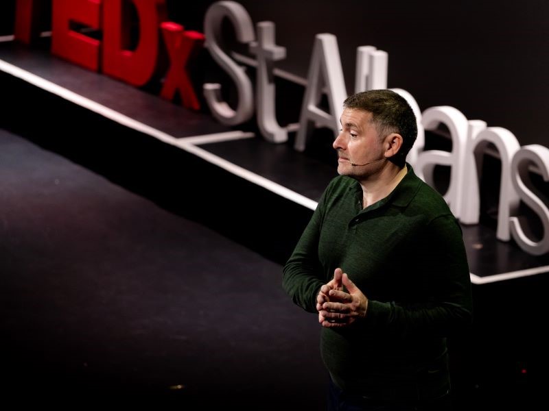 A man in a black shirt stands on a black stage next to a Tedx St Albans sign, giving a talk to an unseen audience.