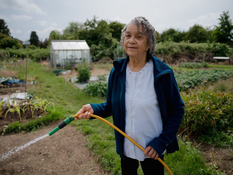 Marion Blake wears a blue open-zip jumper over a white top and holds a hose as she waters her veg patch at her allotment with other veg patches in the background.