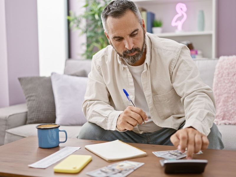 Man sitting on a sofa, checking a shopping list and typing on a calculator.