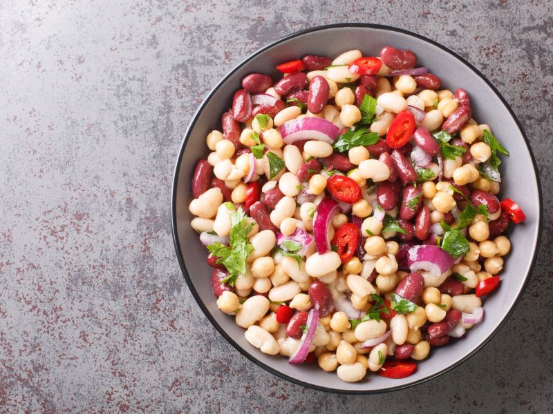 Top view of a bowl of mixed bean salad with tomato, onion and herbs on a grey worktop.