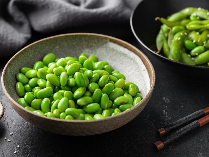 Close up of two bowls of edamame beans, on a black tablecloth with chopsticks just visible.
