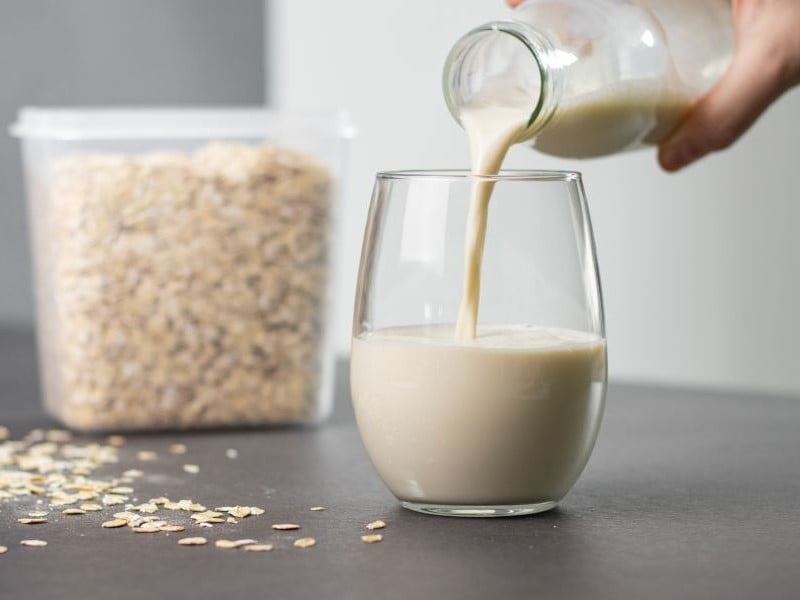 A glass sits on a grey worktop and oat milk is poured into it from a glass bottle. There is a plastic container full of oats in the background.