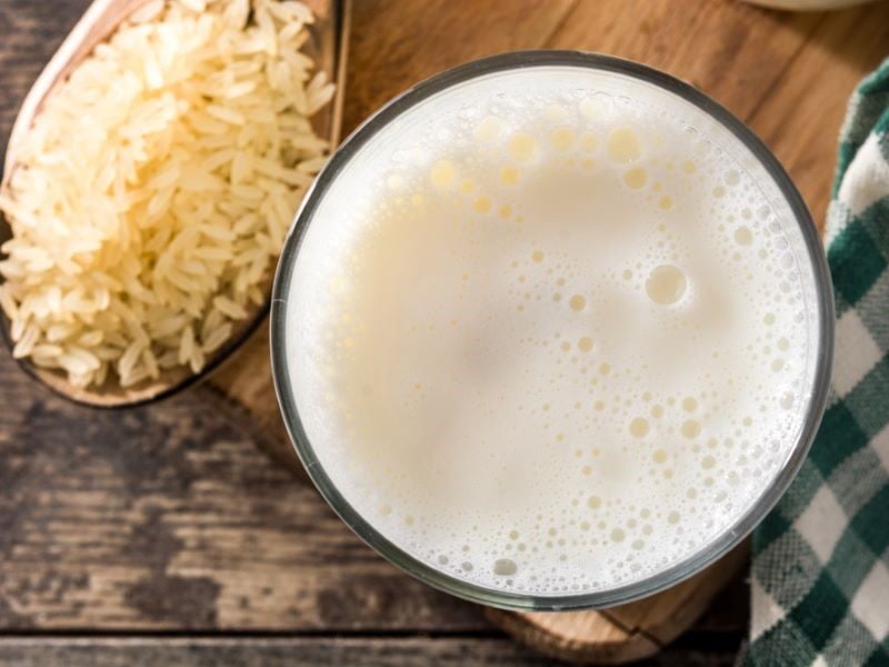 An overhead view of a glass full of rice milk. The glass is stood on a wooden board with a green and white checked tablecloth to the right and a large spoon full of rice to the left.