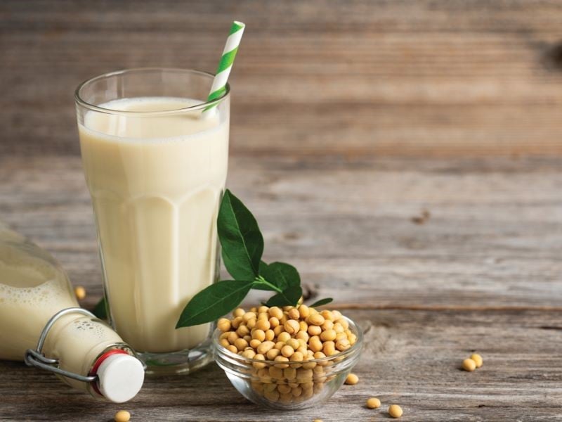 A clear glass full of soya milk, with a green and white stripy straw. There is a small bowl of soy beans to the right and a fallen-over glass bottle of soya milk lying to the left.