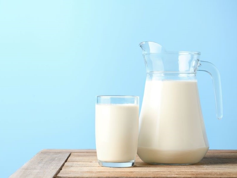 A glass jug of cream-coloured milk sits on a wooden table next to a glass of milk, with a light blue backdrop.