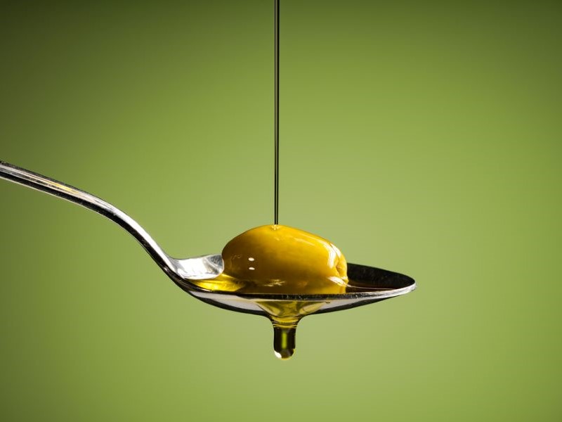 A sliver spoon is suspended mid-air, sideways on, against an olive green background. There is a green olive on the spoon and olive oil being drizzled onto it.