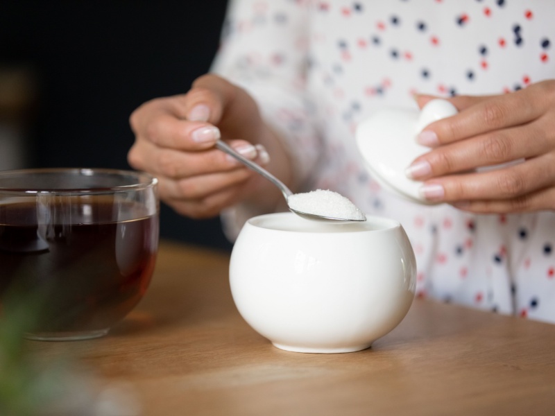 A woman wearing a white spotty shirt holds a teaspoon of sugar over a white sugar bowl. A see-through cup of black tea is also on the table.