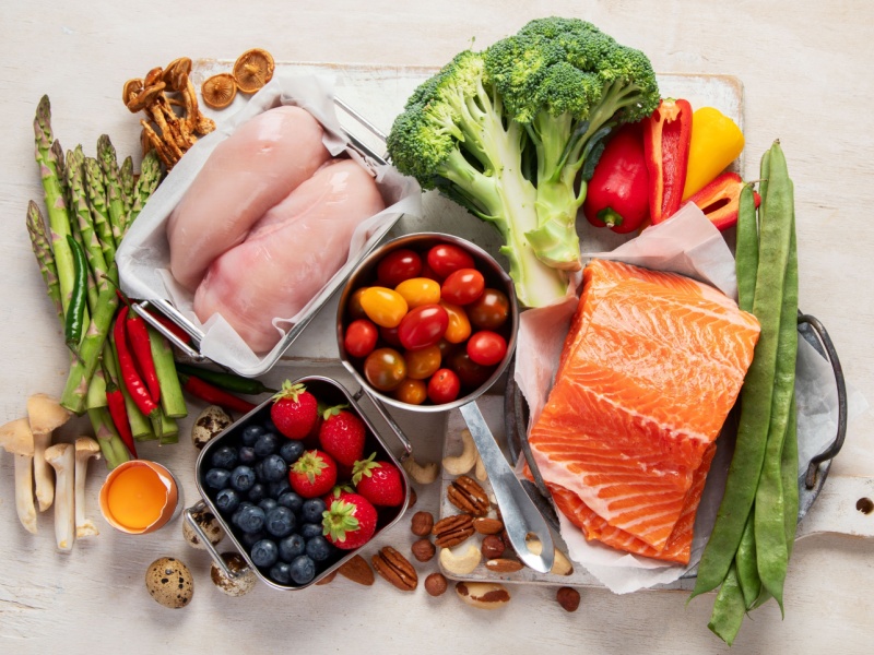 An overhead image of healthy foods including raw salmon and chicken breast, with fruit, vegetables and nuts.