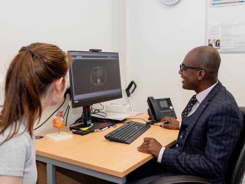 Professor Akowuah in a suit and tie consulting with a heart patient and looking at a scan image on a monitor, at The James cook University Hospital, Middlesbrough.