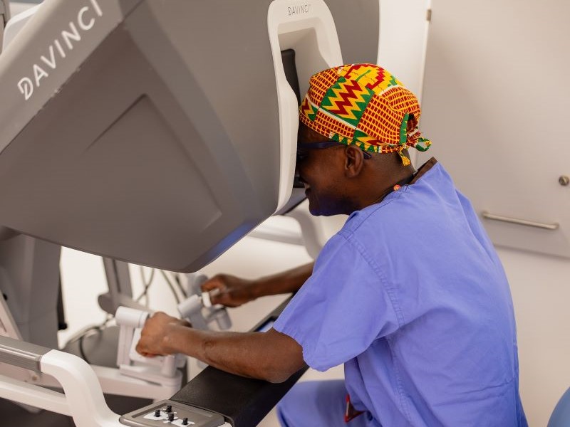 Professor Akowuah in blue scrubs and a colourful head cover sitting at the console of a Da Vinci surgical robot in theatre at The James Cook University Hospital, Middlesbrough.