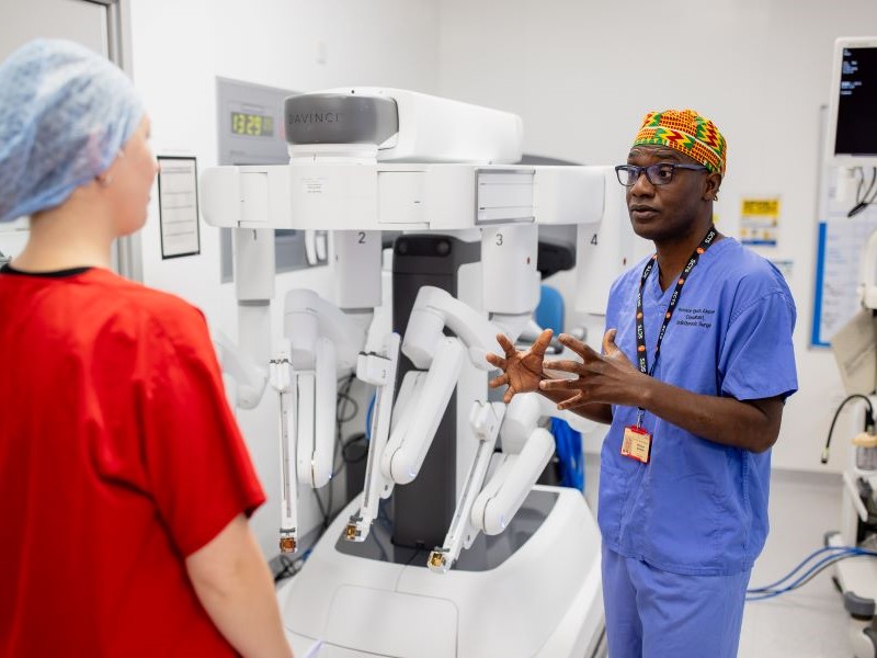 Professor Akowuah in blue scrubs talking with a colleague in red scrubs and standing next to a Da Vinci surgical robot, at The James Cook University Hospital in Middlesbrough.