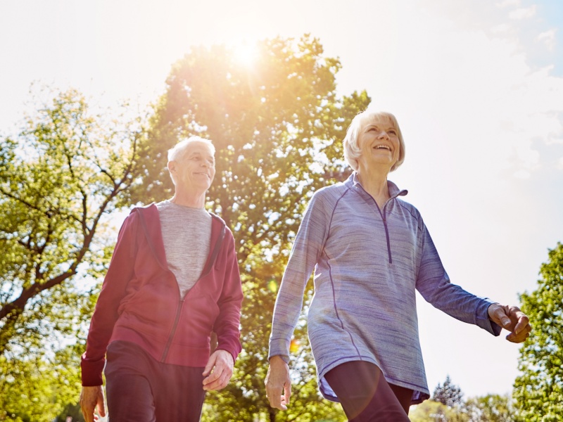A senior couple wearing sports clothing walking outside on a sunny day with trees in the background.