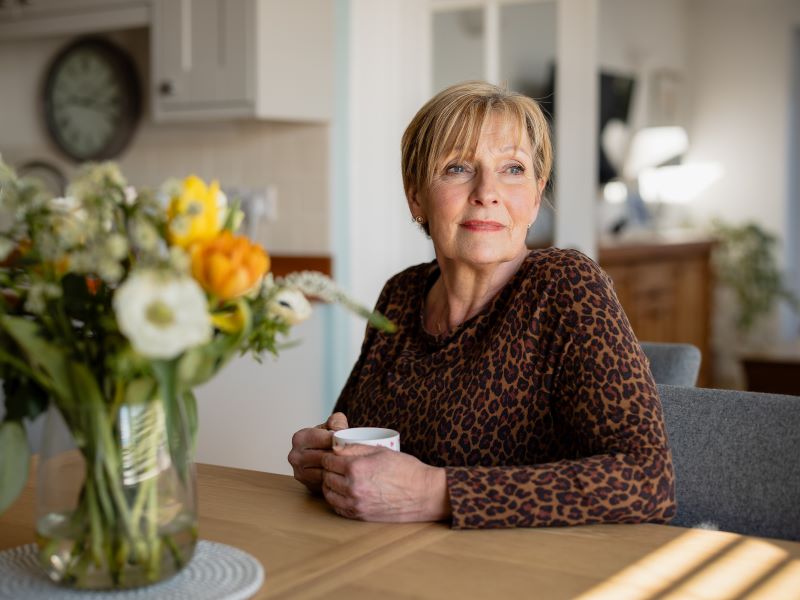 Linda Watling sitting at her kitchen table with flowers holding a mug of tea.