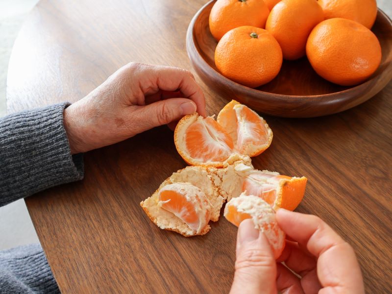Close up of a senior person's hand with a peeled satsuma and a bowl of satsumas on a wooden table.