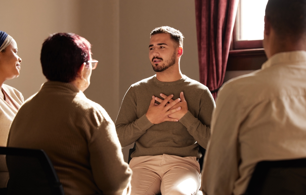 A small circle of people of various ages and backgrounds sitting and talking, with the focus on a male holding his hands to his chest
