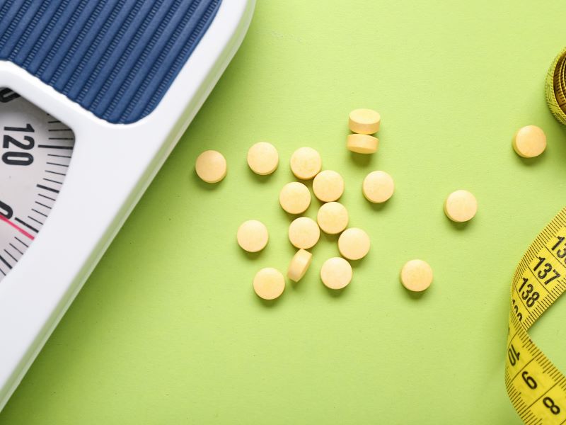 Pills on a green table next to weighing scales and a tape measure.