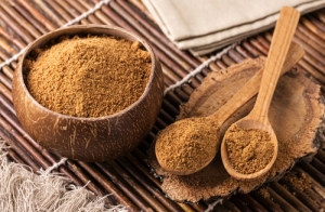 brown coconut palm sugar in a coconut bowl with wooden spoons full of coconut palm sugar resting beside the bowl, all on a brown wooden table mat