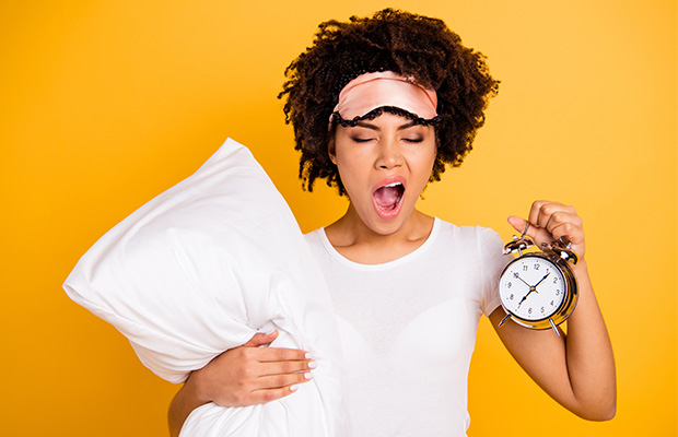 A woman yawning holding a pillow and alarm clock.