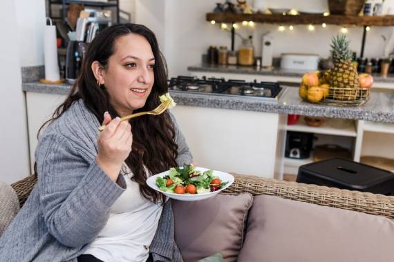 a woman wearing a grey cardigan sitting on a grey sofa and smiling as she holds a bowl of salad in her left hand