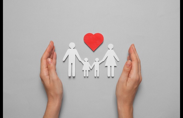 The silhouette of a family holding hands, made out of white paper. A red heart sits above them, also made of paper. A pair of hands frames the scene.