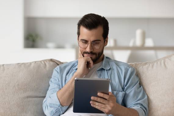 a man with brown hair and a smart blue shirt sitting on a grey sofa smiling and staring down at his tablet computer