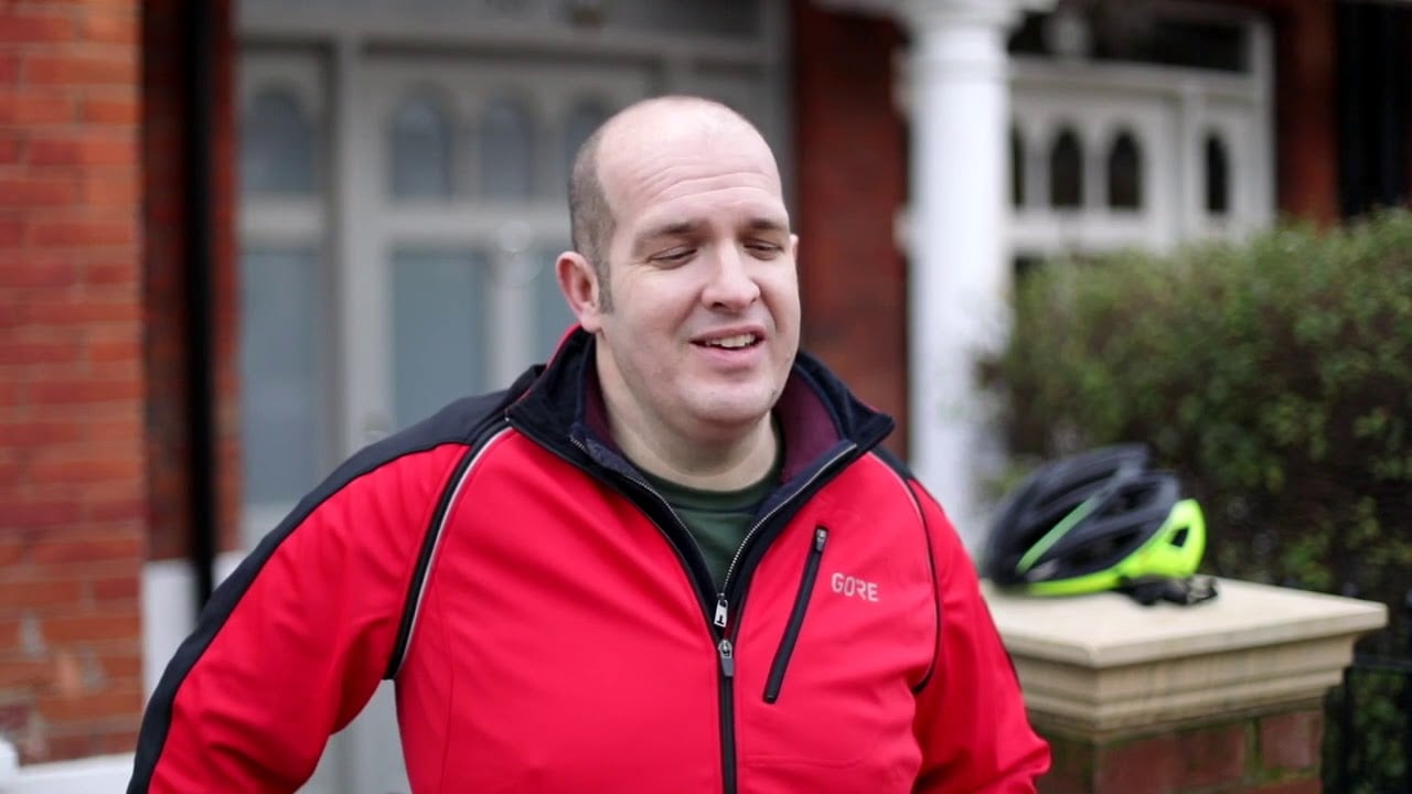Photo of a bald man in a red zip up jumper standing in front of a house