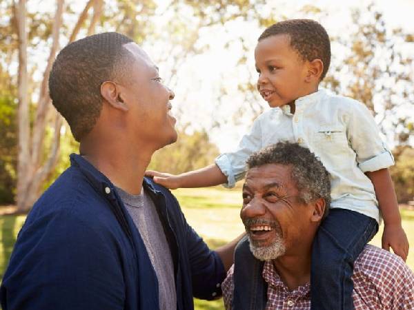 three generations of family including a young father, a toddler and a grandfather stood in a park and smiling at each other