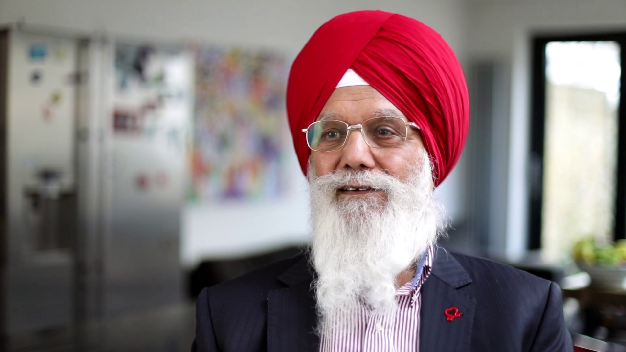 Man with a white beard and moustache wearing pale glasses and a red turban, He is wearing a purple shirt and dark blazer, sat looking at the camera.