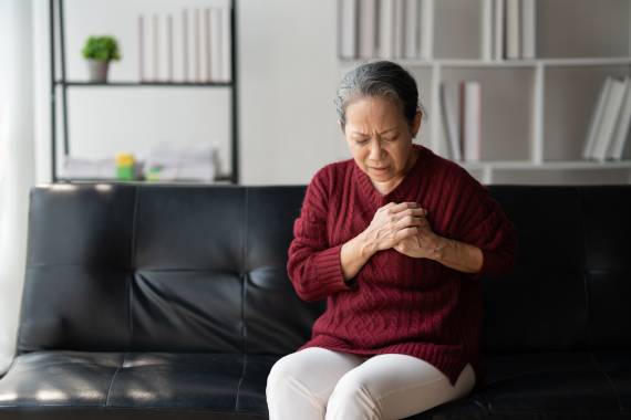 an older chinese woman wearing a long sleeved red shirt and sitting on a black sofa she is clutching the left side of her chest in pain