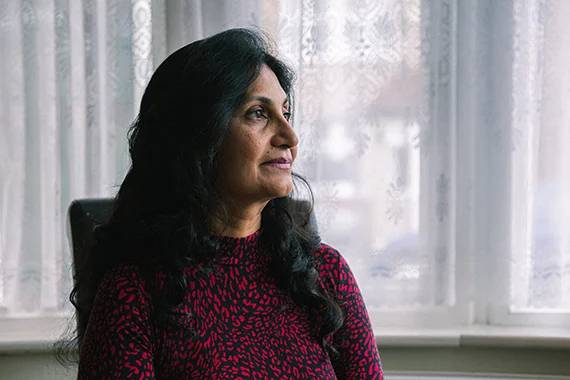 a woman of South Asian heritage sitting in a slightly darkened room and smiling as she stares out of her window