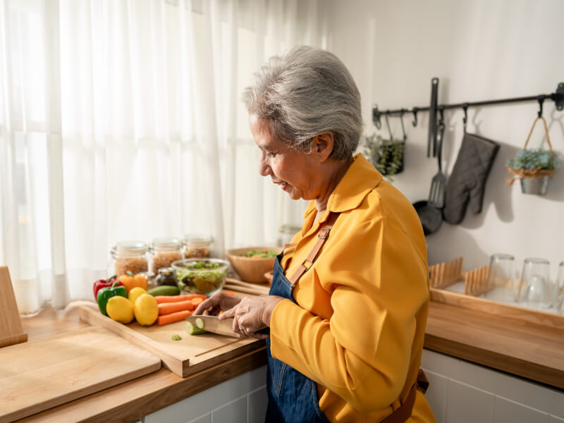 Woman wearing yellow shirt at kitchen counter chopping vegetables. 