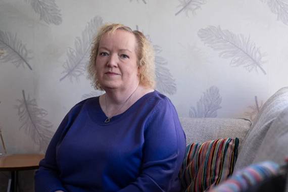 a white older woman in a dark blue long sleeved shirt sitting on a sofa and smiling slightly at the camera