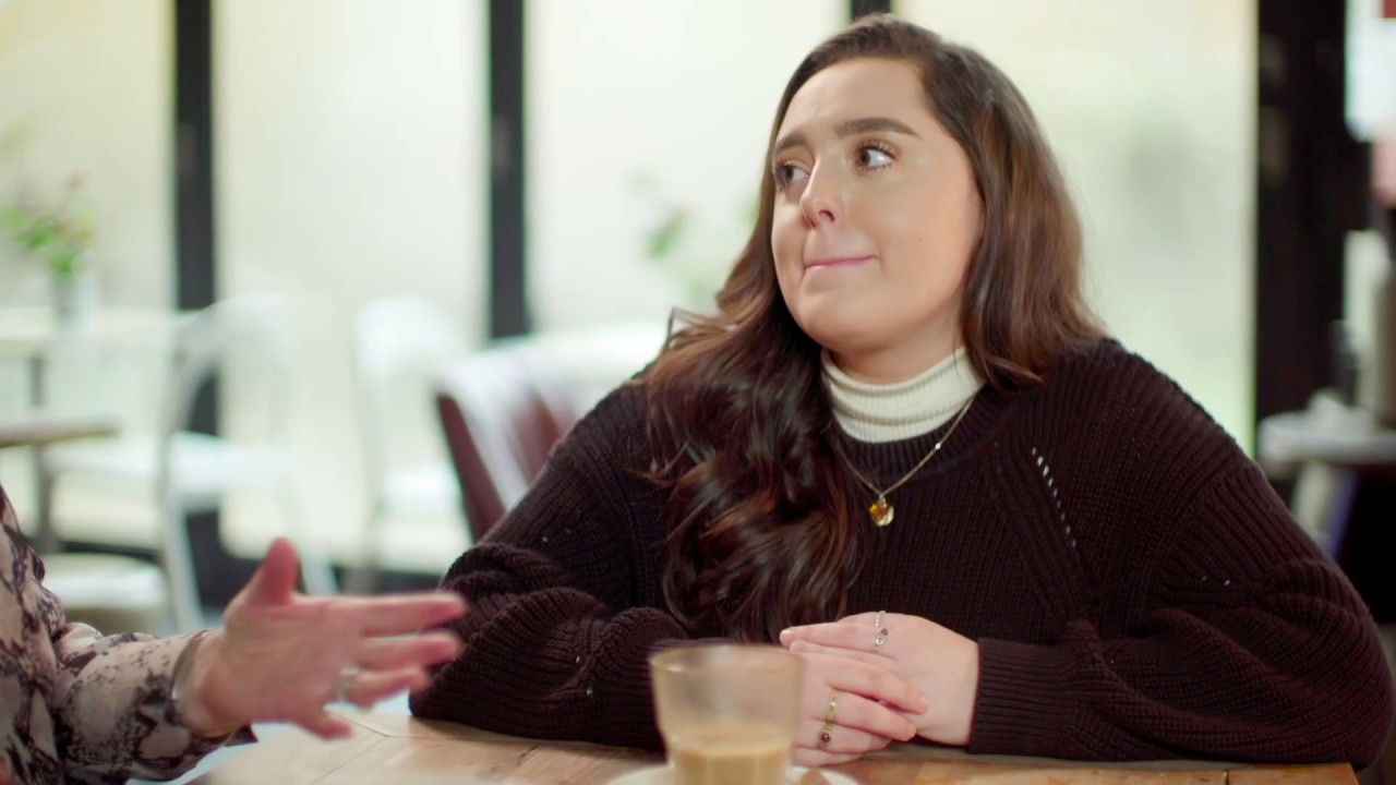 Woman with fair skin and long dark hair sat down at a table, wearing a dark jumper and gold necklace